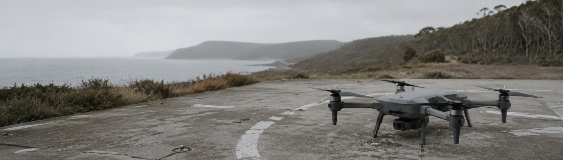 A matte-grey autonomous quadcopter resting on a weathered concrete landing pad at a remote coastal test range, overcast morning.